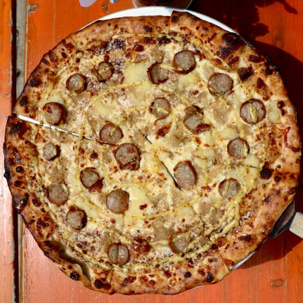 Dieci Pizza's sausage and potato pizza, with a drizzle of mustard, on an orange wooden table.